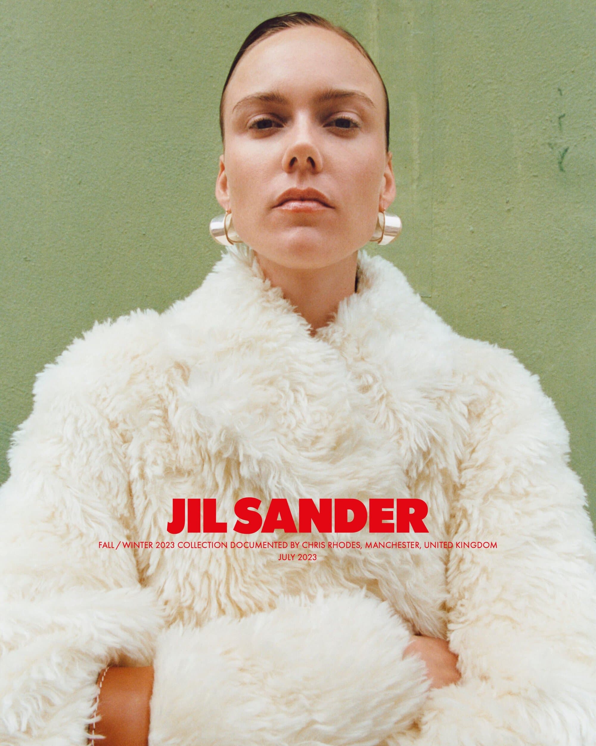 Blonde woman with neutral expression wearing large silver earrings and a white fur coat against an olive green background.