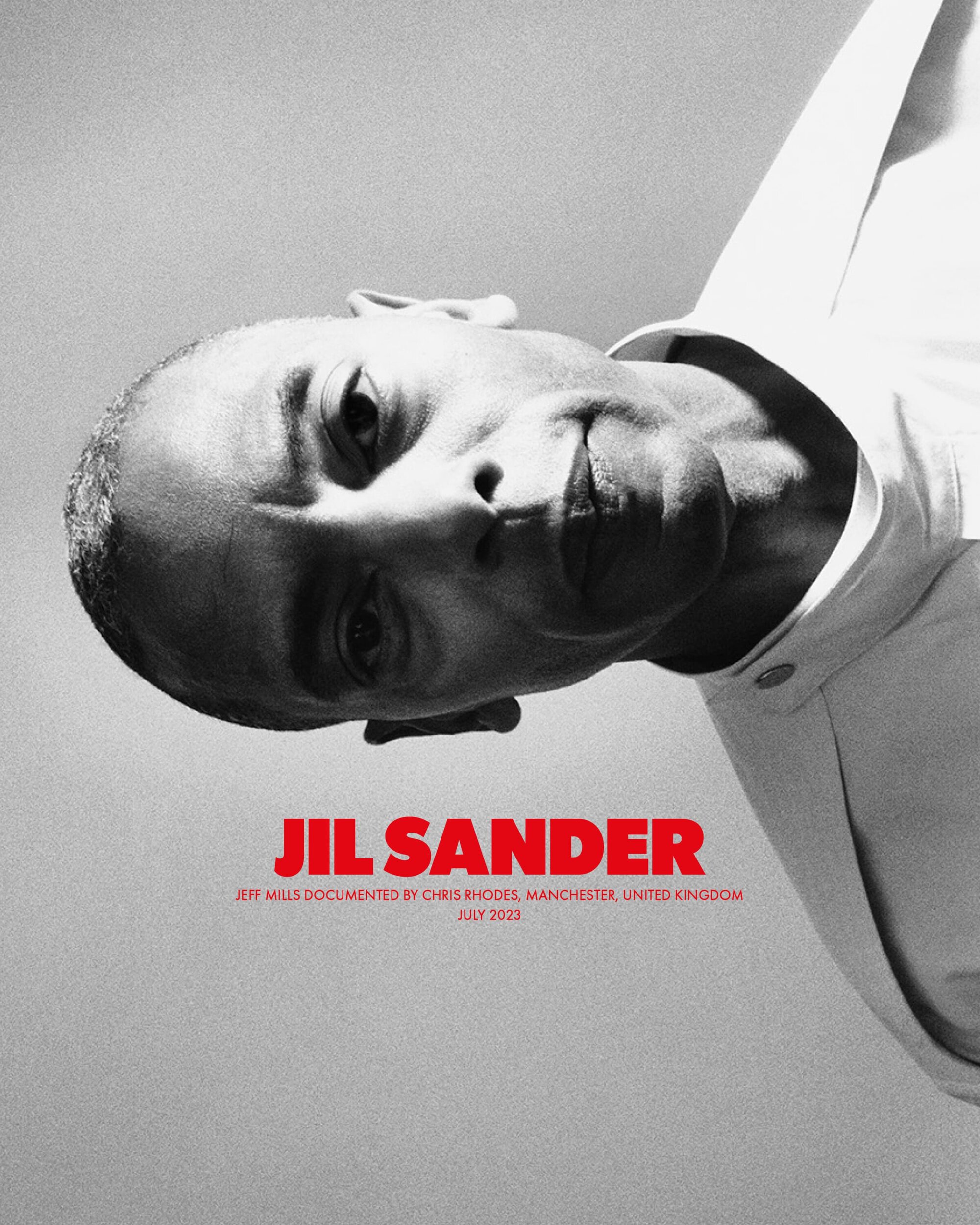 Close-up black and white portrait of a bald adult male wearing a white shirt, angled slightly downward, with Jil Sander text overlay.