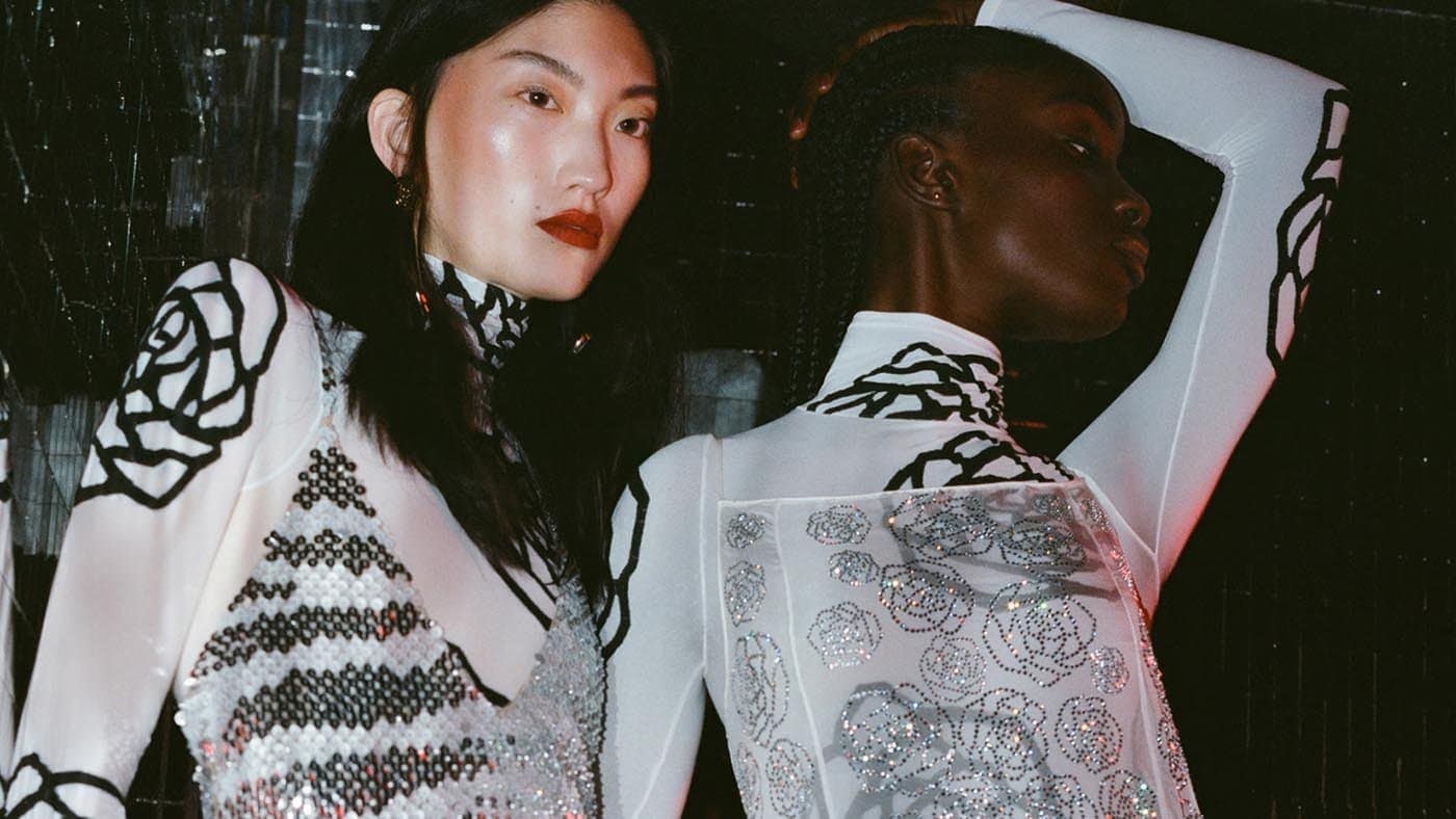 Two fashion models pose indoors, wearing white dresses with black and silver floral designs, showcasing stylish and modern apparel.