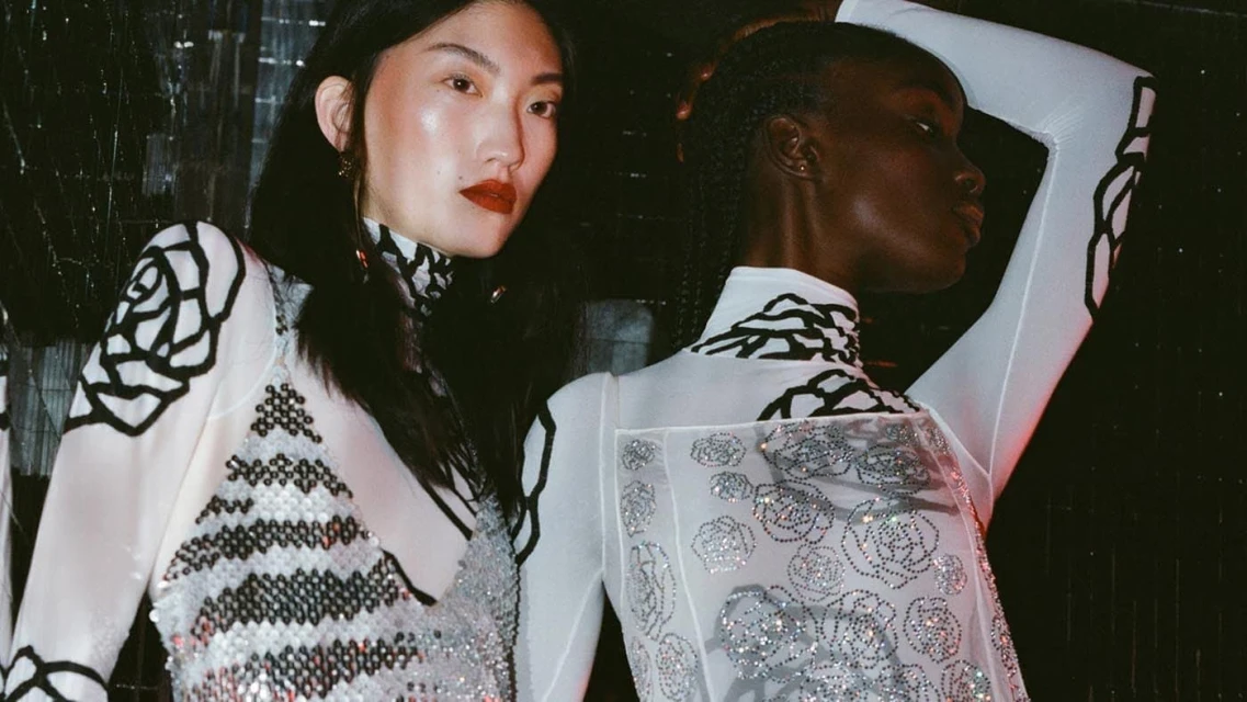 Two fashion models pose indoors, wearing white dresses with black and silver floral designs, showcasing stylish and modern apparel.