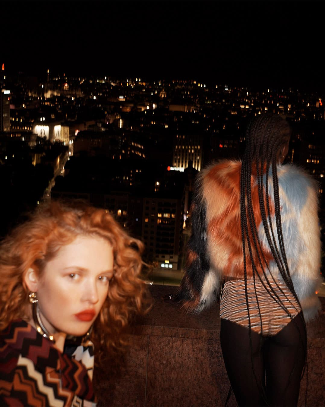 Two women pose on a rooftop at night with city lights in the background, featuring colorful fur coats and patterned clothing.