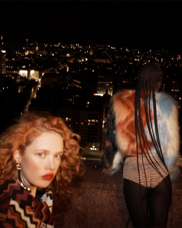 Two women pose on a rooftop at night with city lights in the background, featuring colorful fur coats and patterned clothing.
