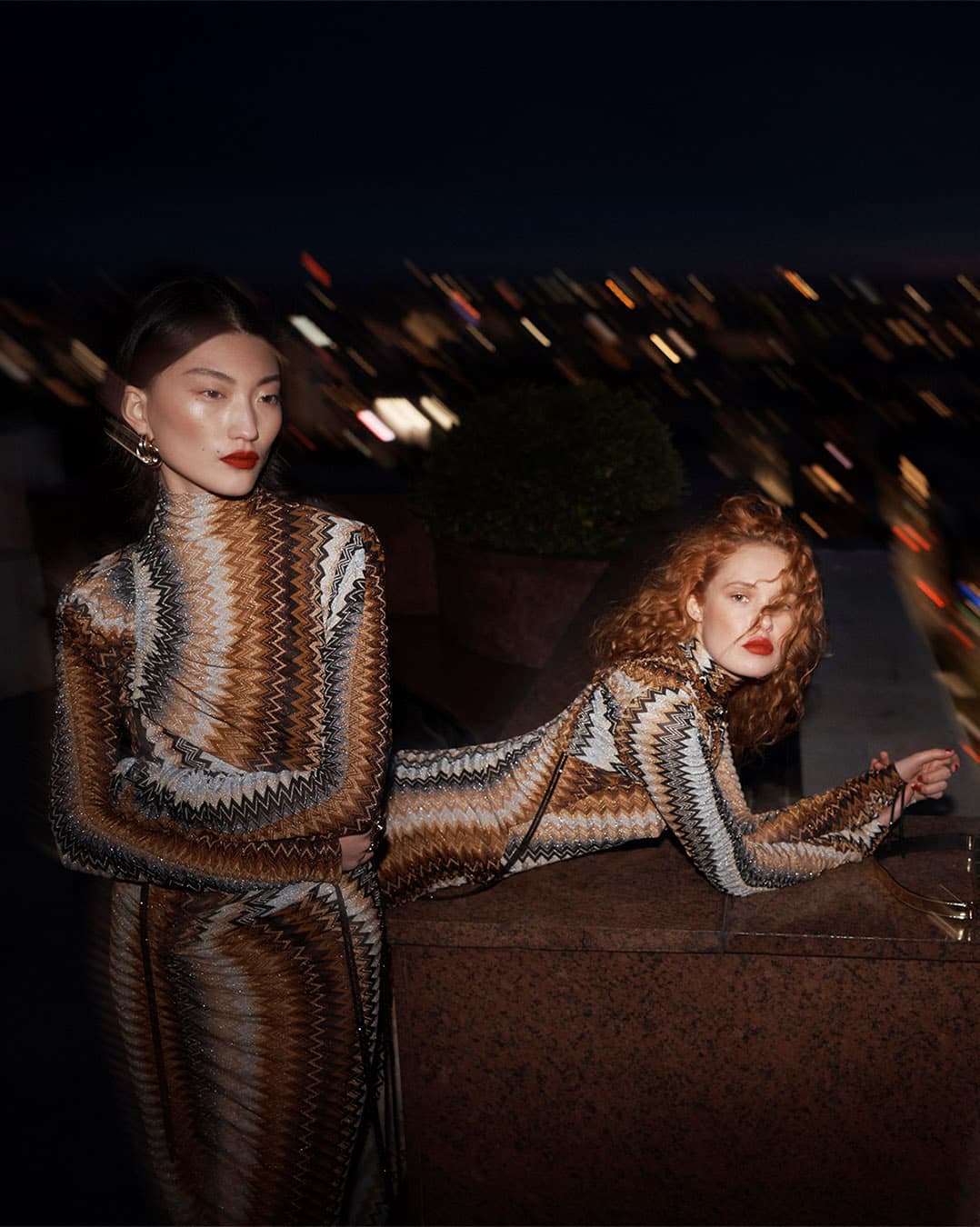 Two fashion models pose in patterned dresses against an out-of-focus city backdrop at night. One model is standing, the other reclines.