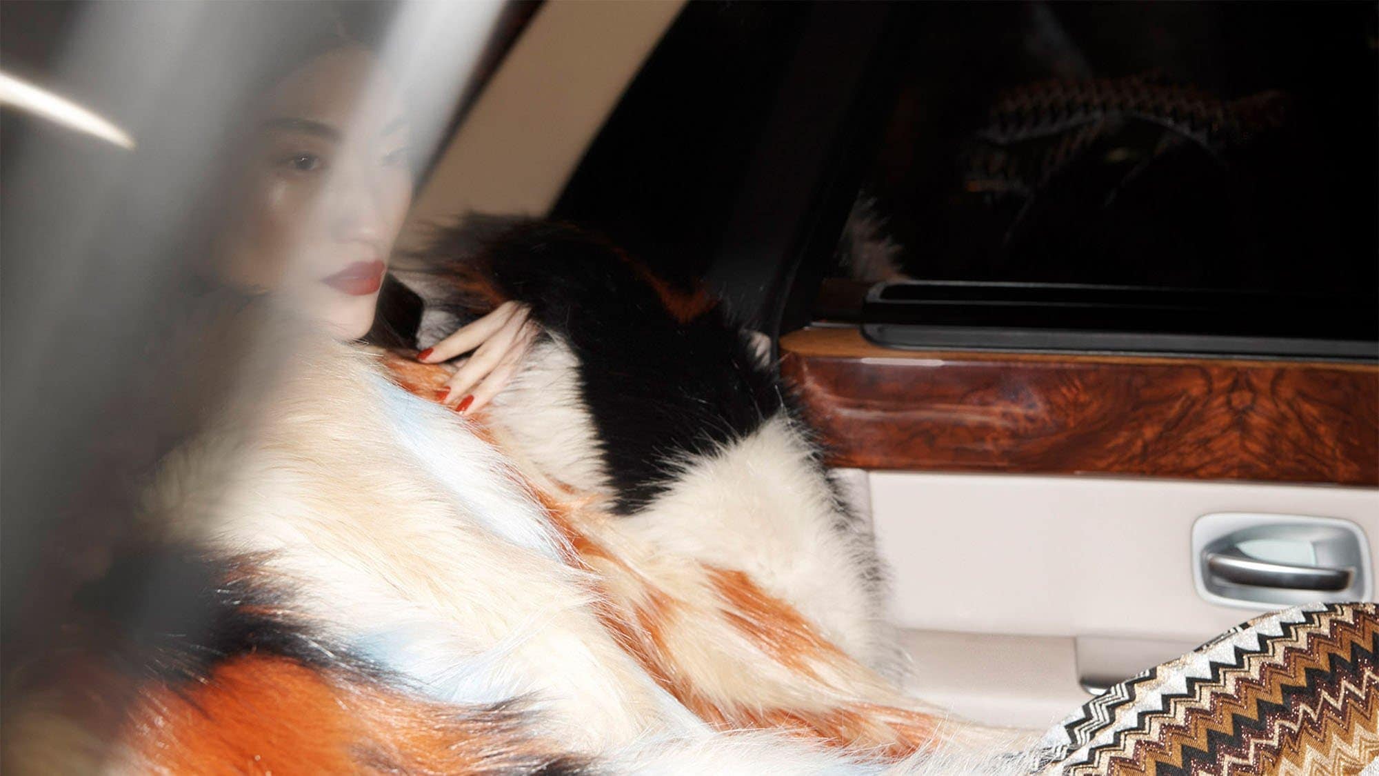 Glamorous woman in colorful fur coat sits inside luxury car, red lipstick and nails, sophisticated style, modern fashion photography.