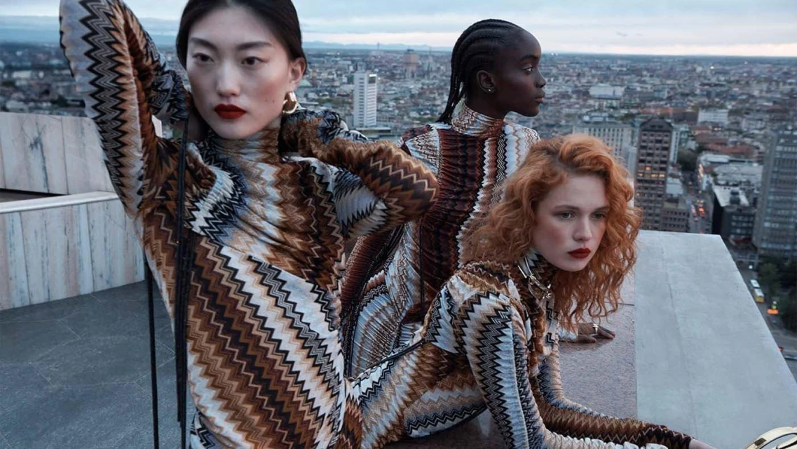 Three fashion models pose on a rooftop in patterned dresses against a cityscape backdrop, featuring a mix of earth-tone colors and modern architecture.