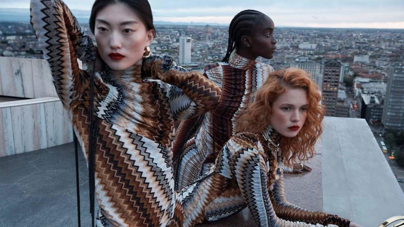Three fashion models pose on a rooftop in patterned dresses against a cityscape backdrop, featuring a mix of earth-tone colors and modern architecture.