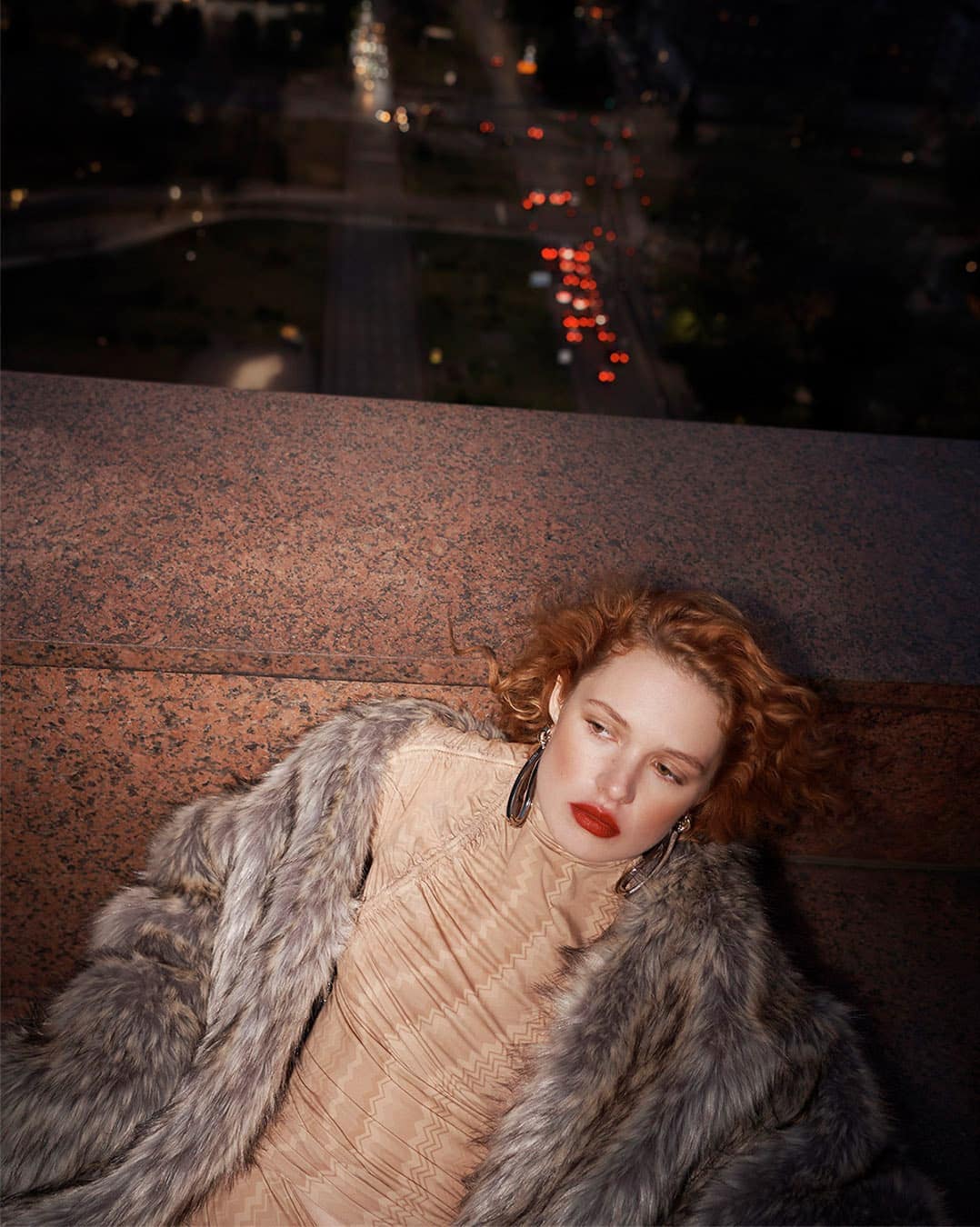 A woman with red hair and red lipstick lies on a stone ledge with a fur coat, with a blurry night cityscape in the background.