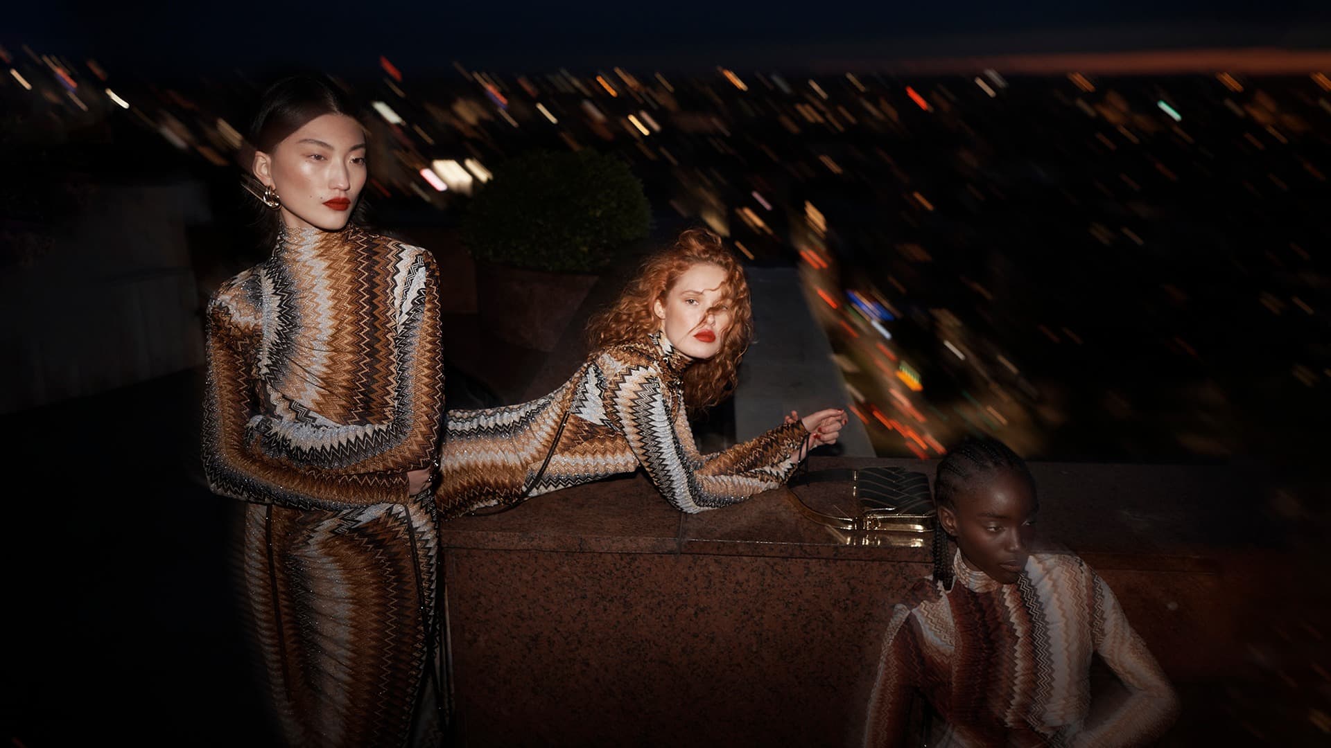 Three fashion models wearing zigzag patterned long-sleeved dresses pose against a city skyline at night with blurred lights in the background.