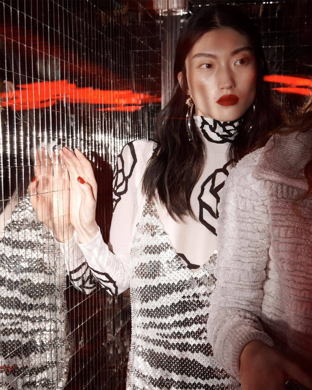 Asian woman in a white floral-patterned dress and long earrings stands next to a reflective, metallic grid surface in a dimly lit room.