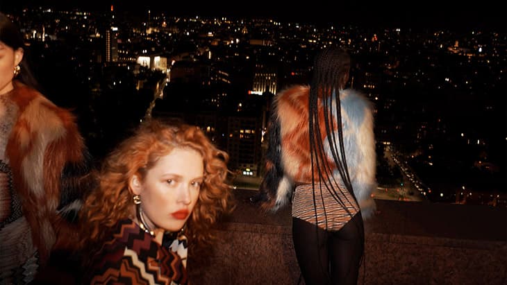 Three fashion models pose against a dark city skyline at night, wearing colorful patterned clothing and faux fur jackets in soft lighting.