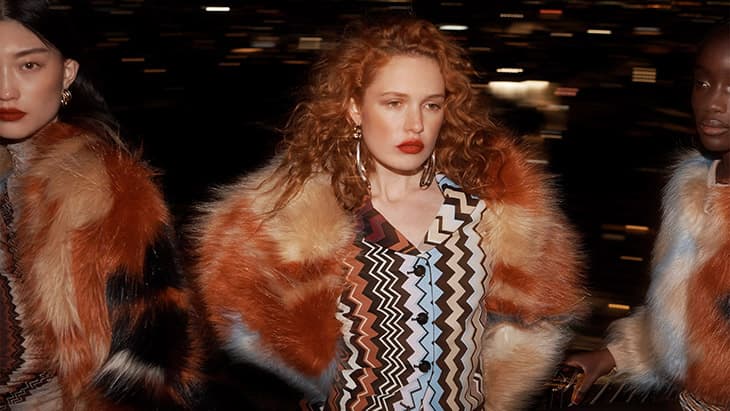 Three women in patterned outfits and fur-trimmed jackets stand against a blurred nighttime cityscape, fashion, style, and urban elegance.