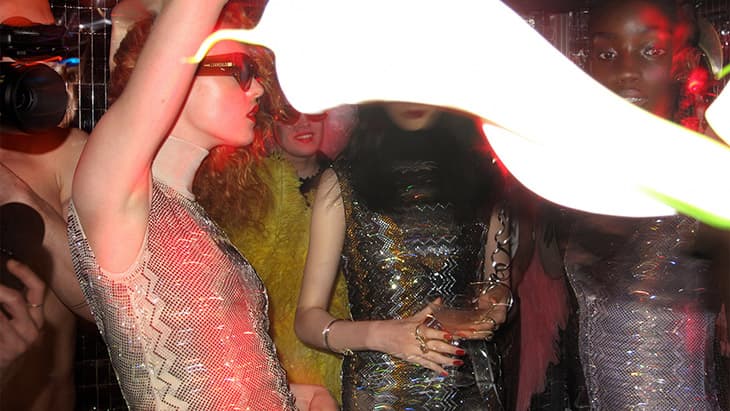 Group of fashionable women pose at a party with strobe lights, wearing sequin dresses and accessories, celebrating in an indoor setting.