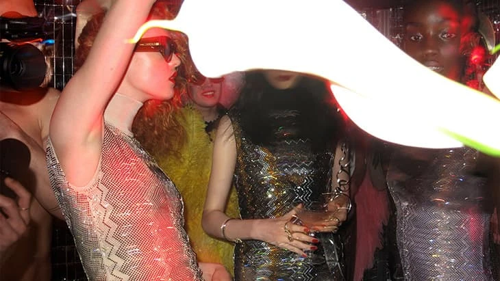 Group of fashionable women pose at a party with strobe lights, wearing sequin dresses and accessories, celebrating in an indoor setting.