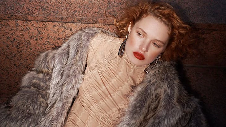 Woman with red hair wearing a fur coat and dress, posing indoors with red lipstick and large earrings, fashion portrait.