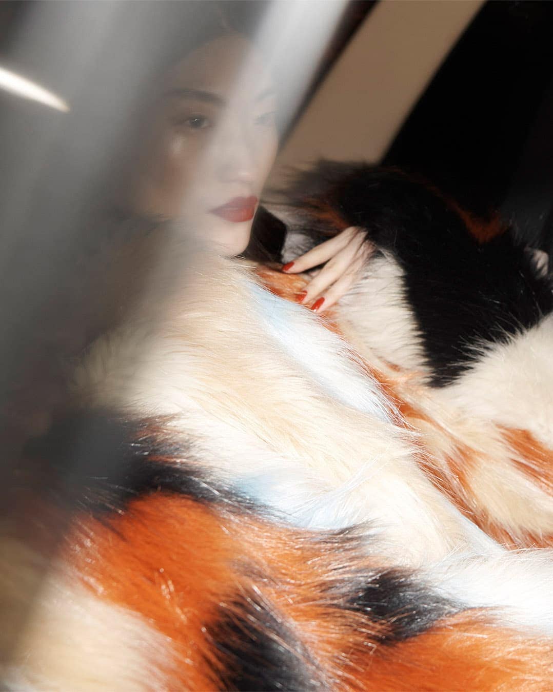 Portrait of a young adult woman with red lipstick and nails, wearing a multi-colored faux fur coat indoors with natural lighting.
