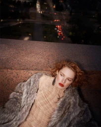 Model with red hair wearing a furry coat leaning against a stone ledge overlooking a blurred night street view