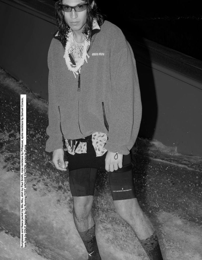 Man in sunglasses and fleece jacket stands in water with white necklace and patterned shorts against dark background in a black and white image.