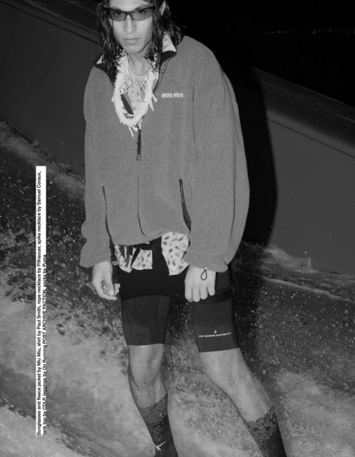 Man in sunglasses and fleece jacket stands in water with white necklace and patterned shorts against dark background in a black and white image.