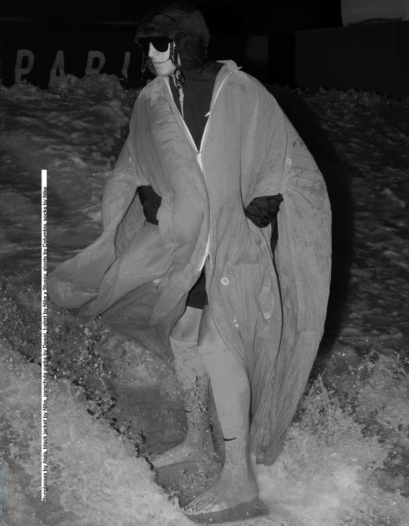 A person in sunglasses and draped clothing stands in shallow water, in a black-and-white photograph with water splashing around the legs.