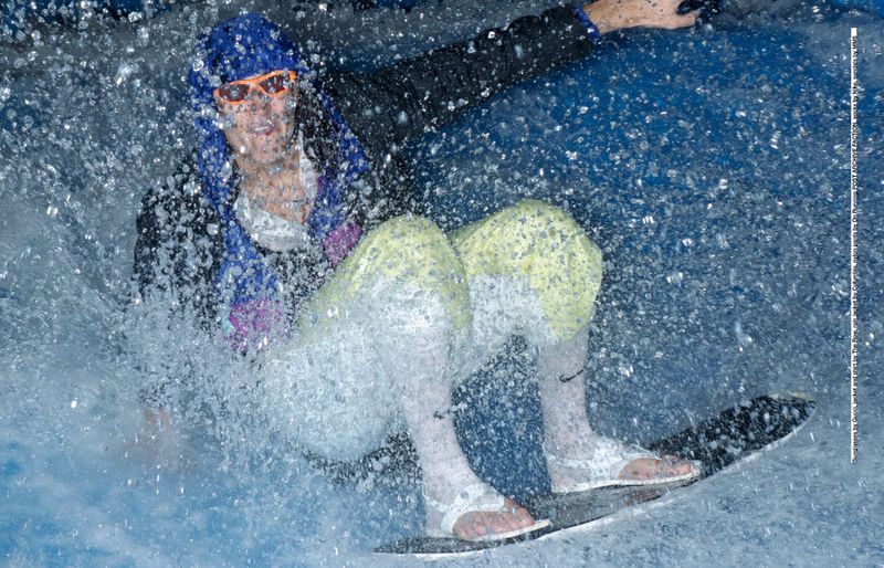 Woman in blue hat and orange goggles rides wave on body board in wetsuit amid splashing water. Water sport activity.