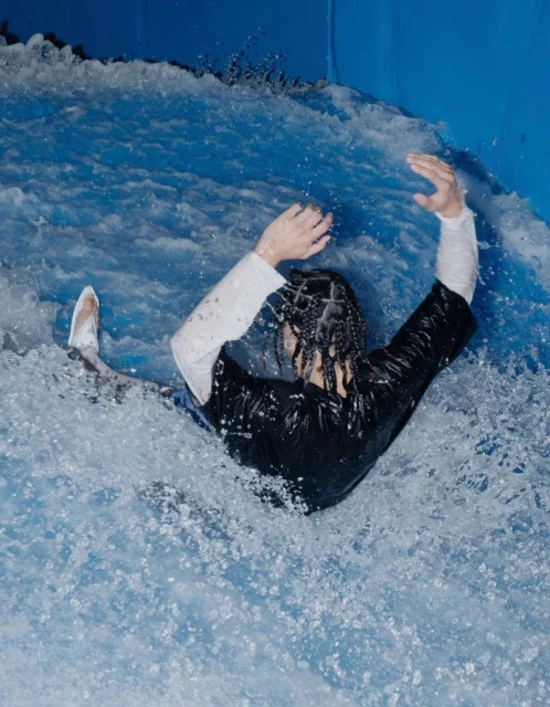 Person in swimwear slides down a wave pool at an indoor water park. The individual wears black and white clothing and braids.