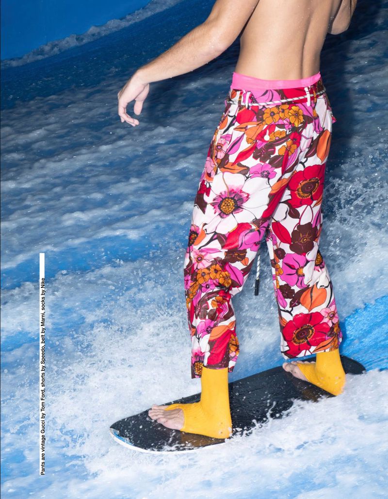 A model surfs indoors wearing floral trousers and yellow socks, captured in a medium shot against a backdrop of artificial wave.