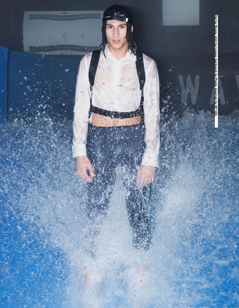 A man in a white shirt and dark pants stands in water, with a backpack, indoor setting, lit by overhead lights, fashion photography.