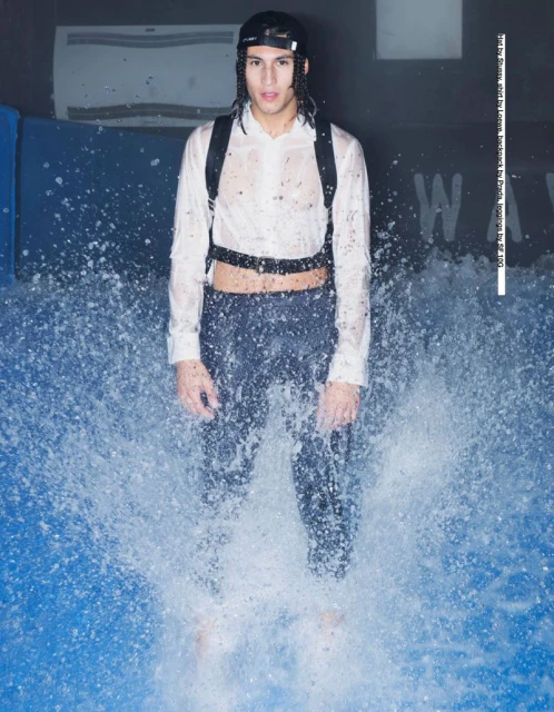 A man in a white shirt and dark pants stands in water, with a backpack, indoor setting, lit by overhead lights, fashion photography.