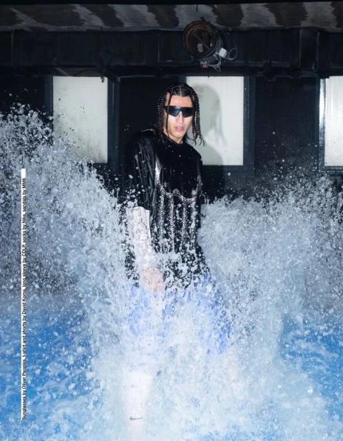 Adult male model stands in a water feature with white splashing water and black industrial architecture in the background.