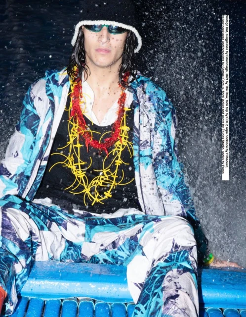 Adult male in a black bucket hat, sunglasses, and patterned jacket sitting in front of water. He wears necklaces in red and yellow.