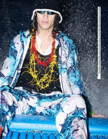 Adult male in a black bucket hat, sunglasses, and patterned jacket sitting in front of water. He wears necklaces in red and yellow.