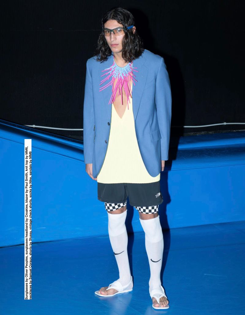 Man in blue blazer, pale yellow top, and black shorts with checkered pattern, accessorized with sunglasses and distinctive necklace.