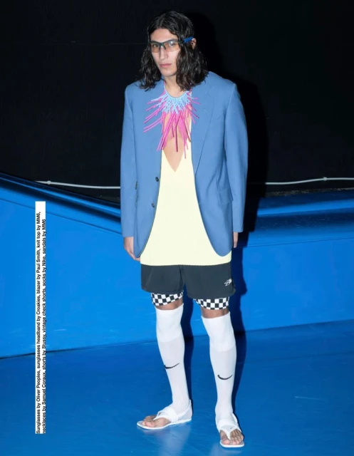Man in blue blazer, pale yellow top, and black shorts with checkered pattern, accessorized with sunglasses and distinctive necklace.