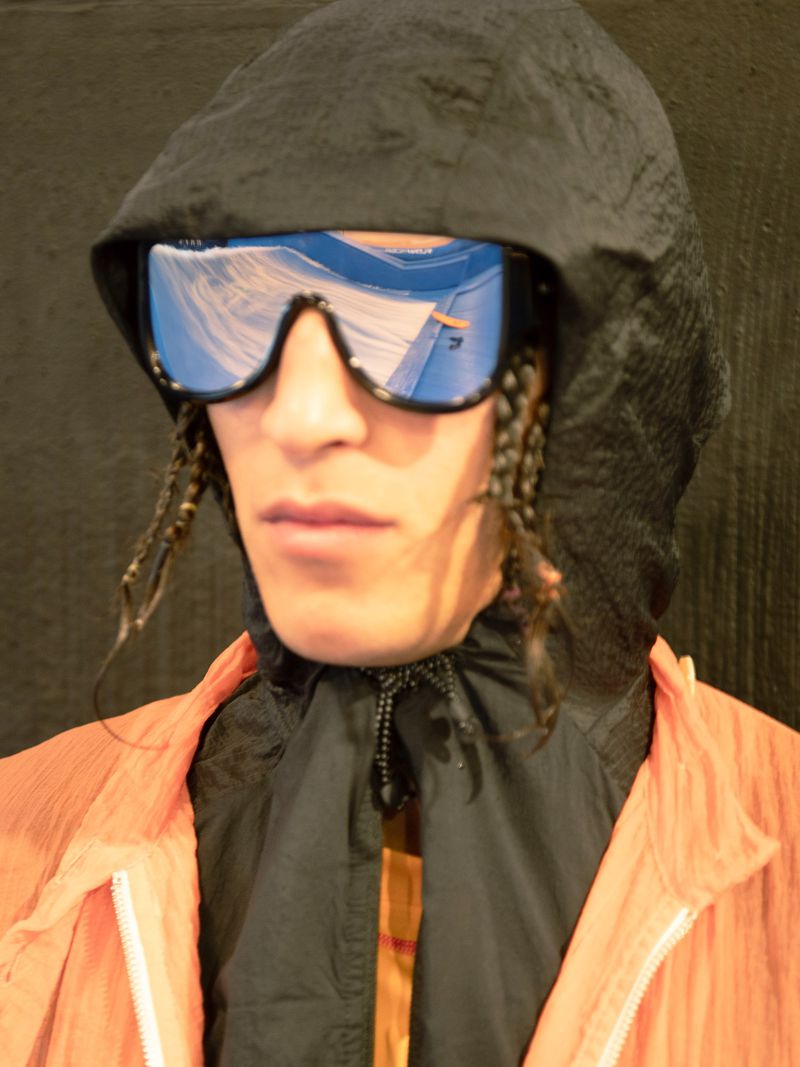 Portrait of an adult male wearing an orange jacket and black hood with reflective blue visor sunglasses, with braided hair.