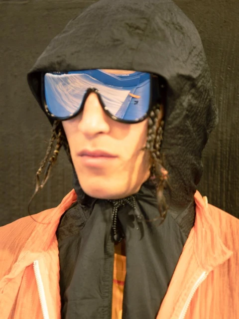 Portrait of an adult male wearing an orange jacket and black hood with reflective blue visor sunglasses, with braided hair.