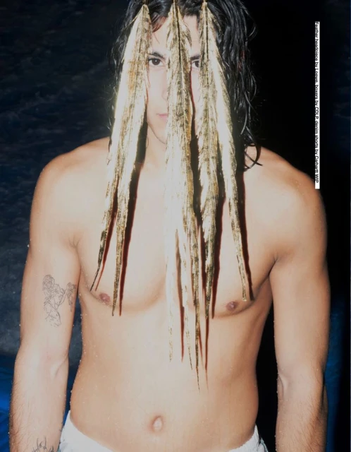 A shirtless person with wet hair stands indoors with feathers in their hair, their face partially obscured by the feathers.