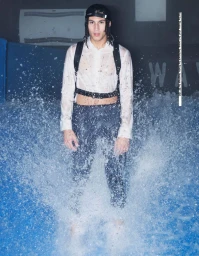 A man standing in water wearing a wet white shirt, dark trousers, and a harness with water splashing around him.