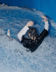 Person sliding through turbulent blue water with arms raised, captured from behind in a high-angle shot.