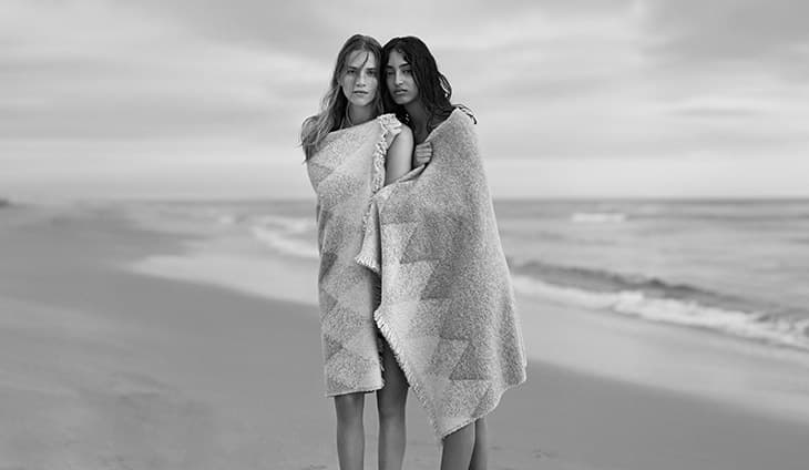 Black and white shot of two women wrapped in patterned blankets on a sandy beach with soft waves and cloudy sky in the background.