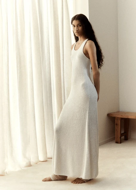 Woman in white dress standing in bright neutral interior setting near window with natural light. Fashion, beauty, and lifestyle concept.