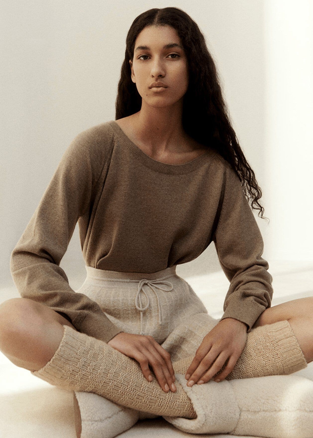Fashion portrait of a woman in a brown top and beige shorts sitting cross-legged against a light background in a studio setting.