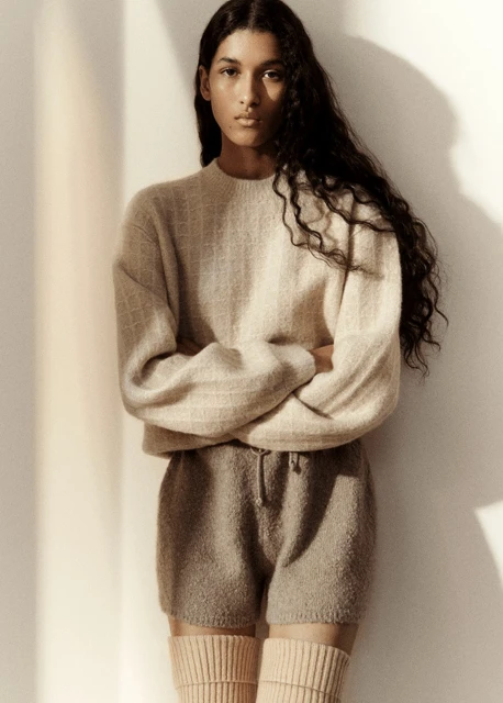 Portrait of a young adult female fashion model with long dark hair, wearing a neutral-toned knit sweater and shorts, studio lit.