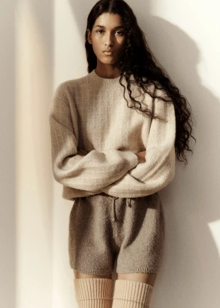 Portrait of a young adult female fashion model with long dark hair, wearing a neutral-toned knit sweater and shorts, studio lit.