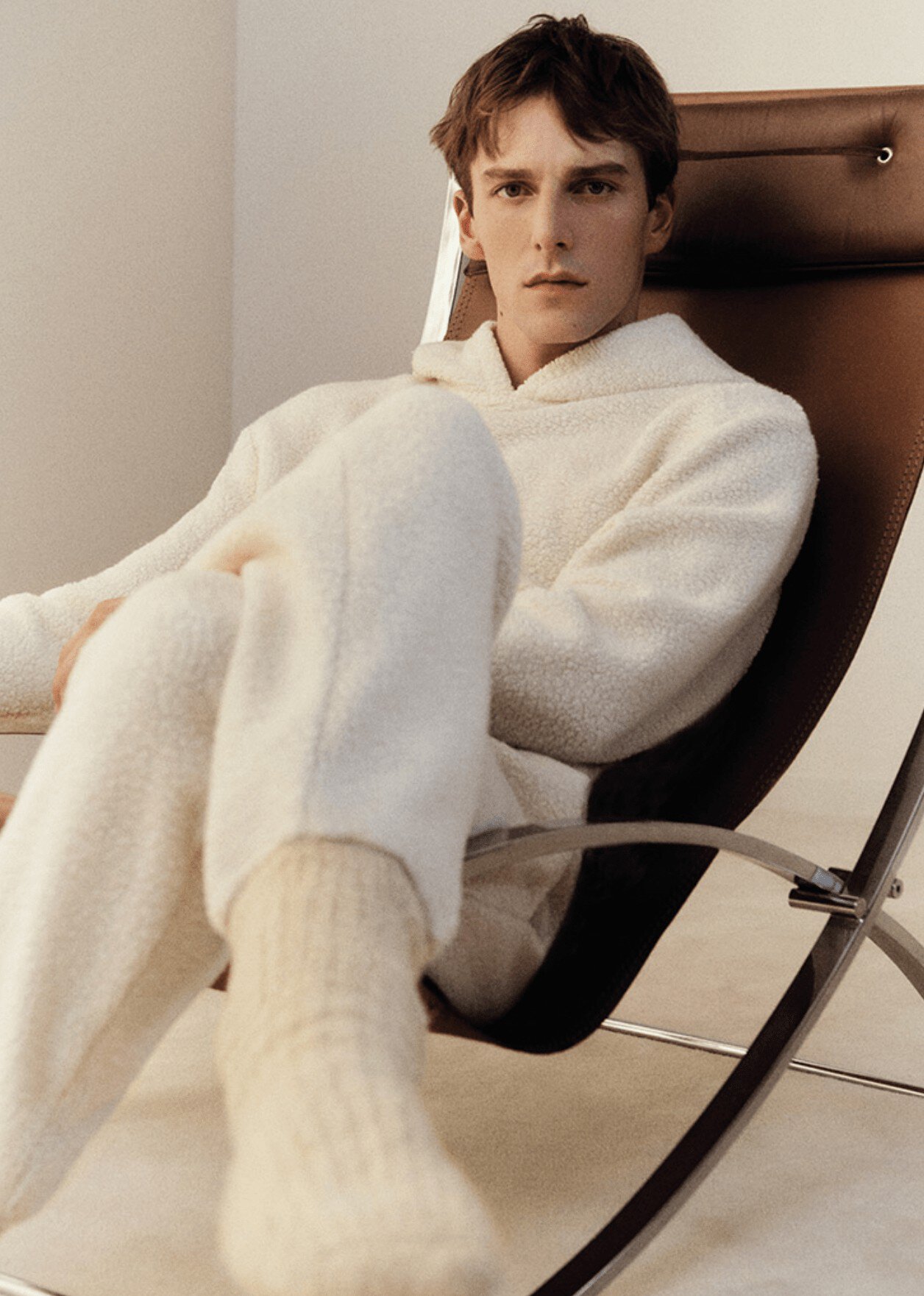 Young adult male model wearing a fuzzy white hoodie and pants is sitting in a modern chair in a neutral studio setting, looking at camera.