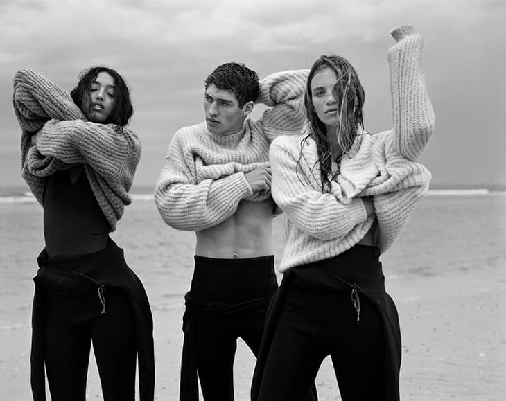 Three people on a beach wearing sweaters and wetsuit pants, posing with arms adjusting their sweaters in a black and white photo.