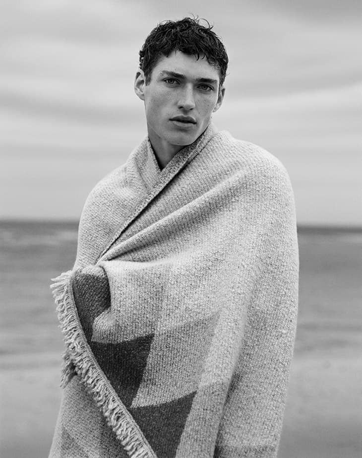 Monochromatic portrait of a young adult male model with dark hair, wrapped in a textured geometric blanket outdoors with a sea backdrop.
