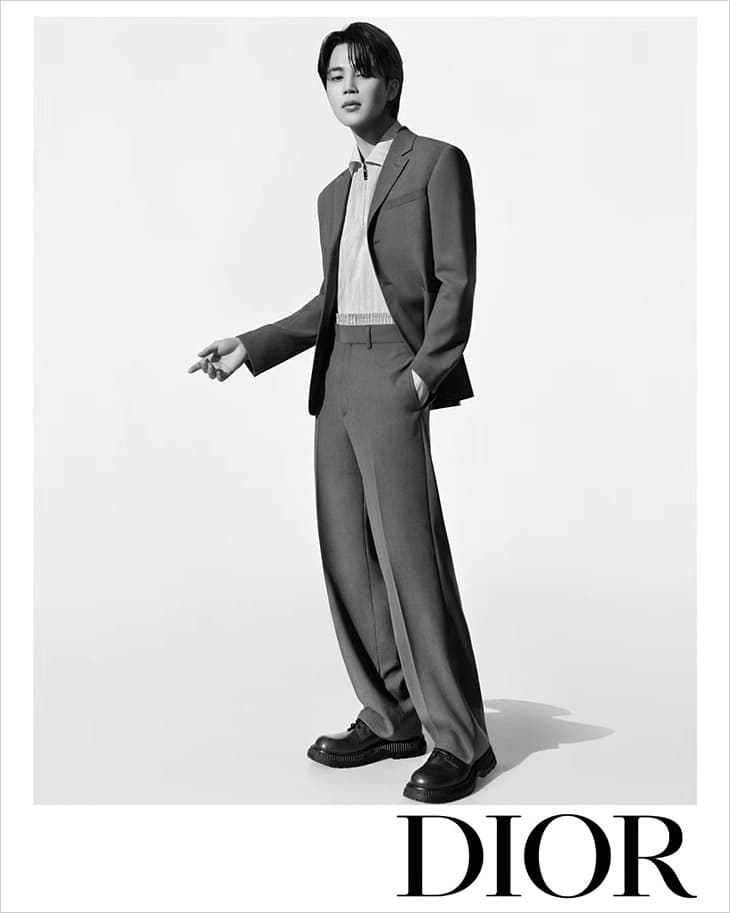 Portrait of a solo adult male model in a grey suit in a photo studio with black shoes against a stark white backdrop; Dior logo visible.