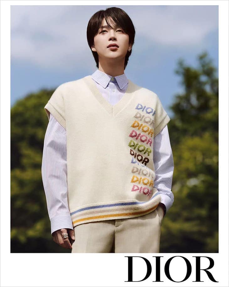 An Asian adult male poses wearing a light blue striped shirt and beige sweater vest with colorful Dior text standing in front of trees.