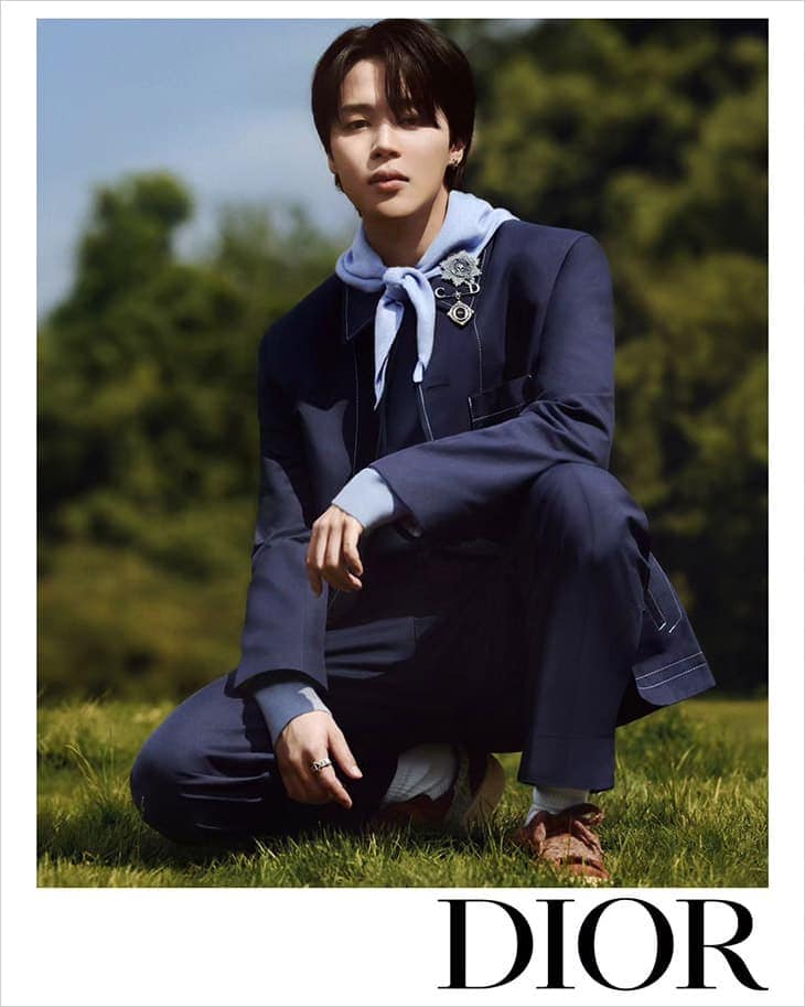 Asian man in a navy suit jacket and light blue hoodie, crouching on a grassy field with a green background, accessorized with rings and brooch.