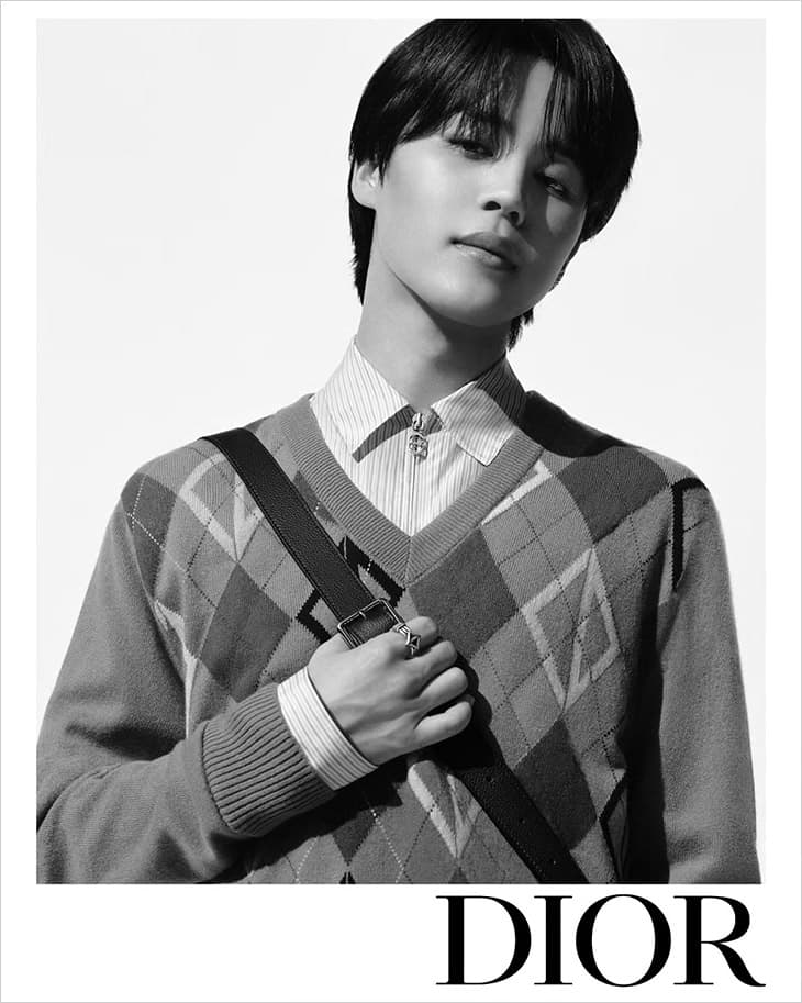 Monochrome portrait of a young adult man in an argyle sweater and collared shirt, accessorized with a shoulder bag and ring. 
